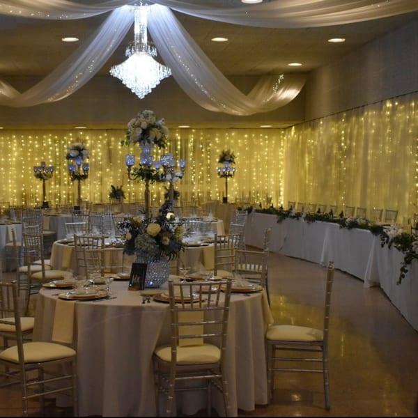 Elegant banquet hall with round tables, white tablecloths, gold chairs, and string lights.