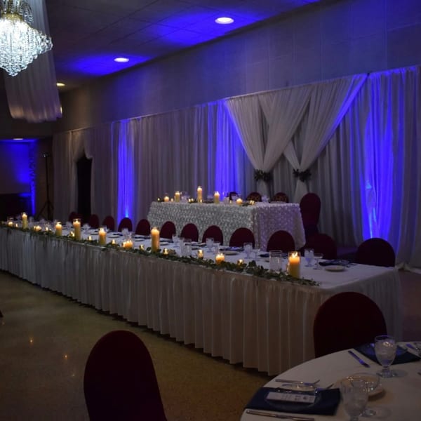 Long dining tables set with candles and place settings, illuminated by blue uplighting and a chandelier.