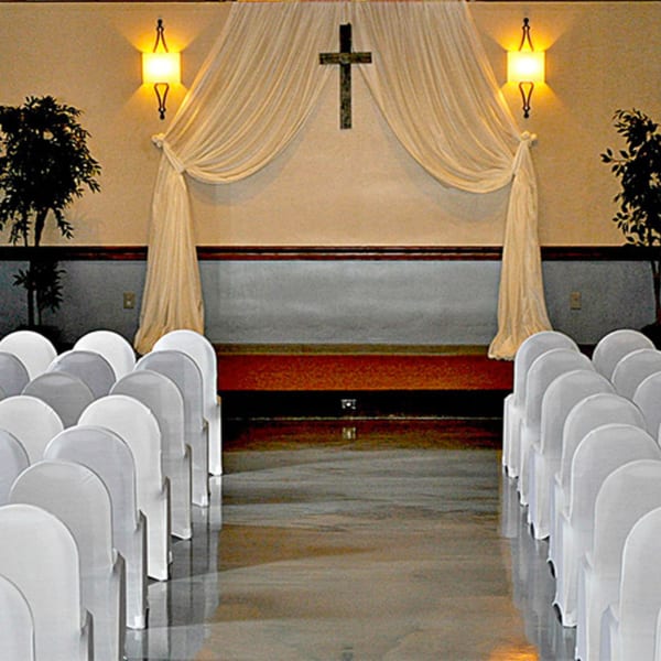 Rows of white covered chairs face a stage with draped fabric, a cross, and wall sconces.