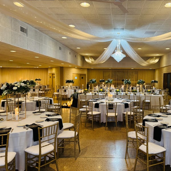 Elegant dining hall with round tables, white tablecloths, black napkins, gold chairs, and floral centerpieces.