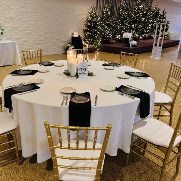 A round table set for a formal event with white tablecloths, black napkins, and candles, surrounded by gold chairs.