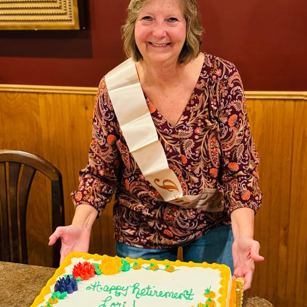 Smiling woman in a patterned shirt and sash holding a white cake with "Happy Retirement Lori!" written on it.