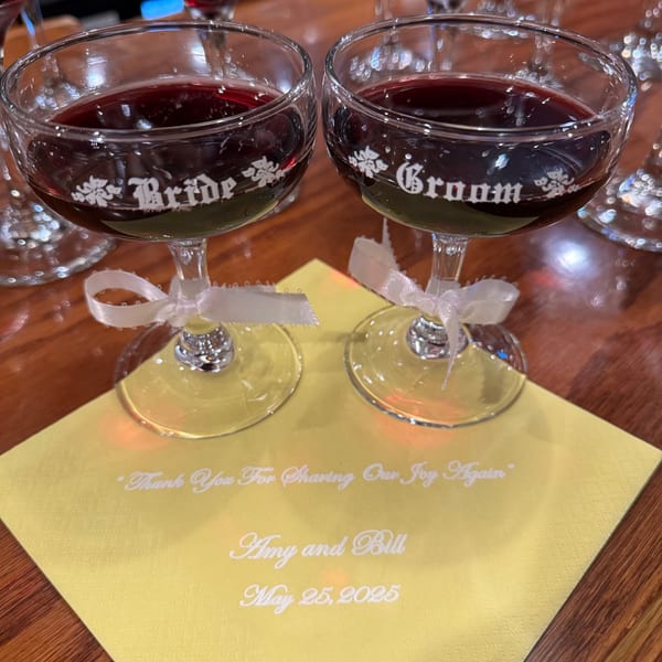Two wine glasses labeled "Bride" and "Groom" with red wine, on a yellow napkin.
