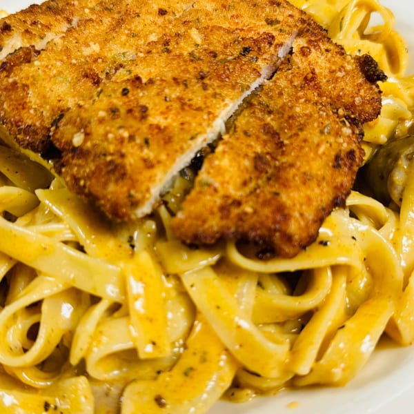 Close-up of golden-brown breaded chicken cutlet, sliced, atop creamy fettuccine pasta.