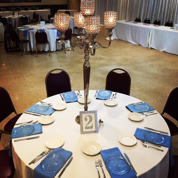 A round table set with blue napkins, white plates, and a candelabra centerpiece in a banquet hall.