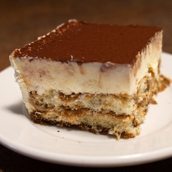 A square slice of tiramisu with layers of cream and cake, topped with cocoa powder.